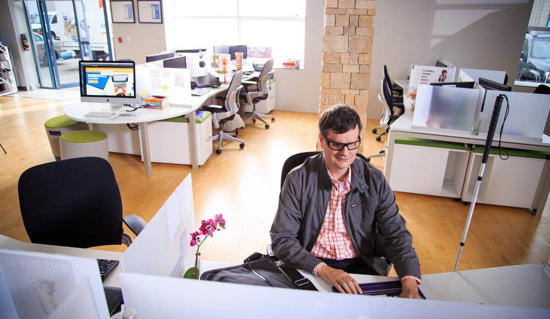 Person with short brown hair wearing a grey jacket working at a computer in an office. There is a white cane by the desk.