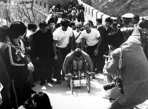 Black and white photo of Rick Hansen surrounded by a crown of people as he pushes up the Great Wall of China.