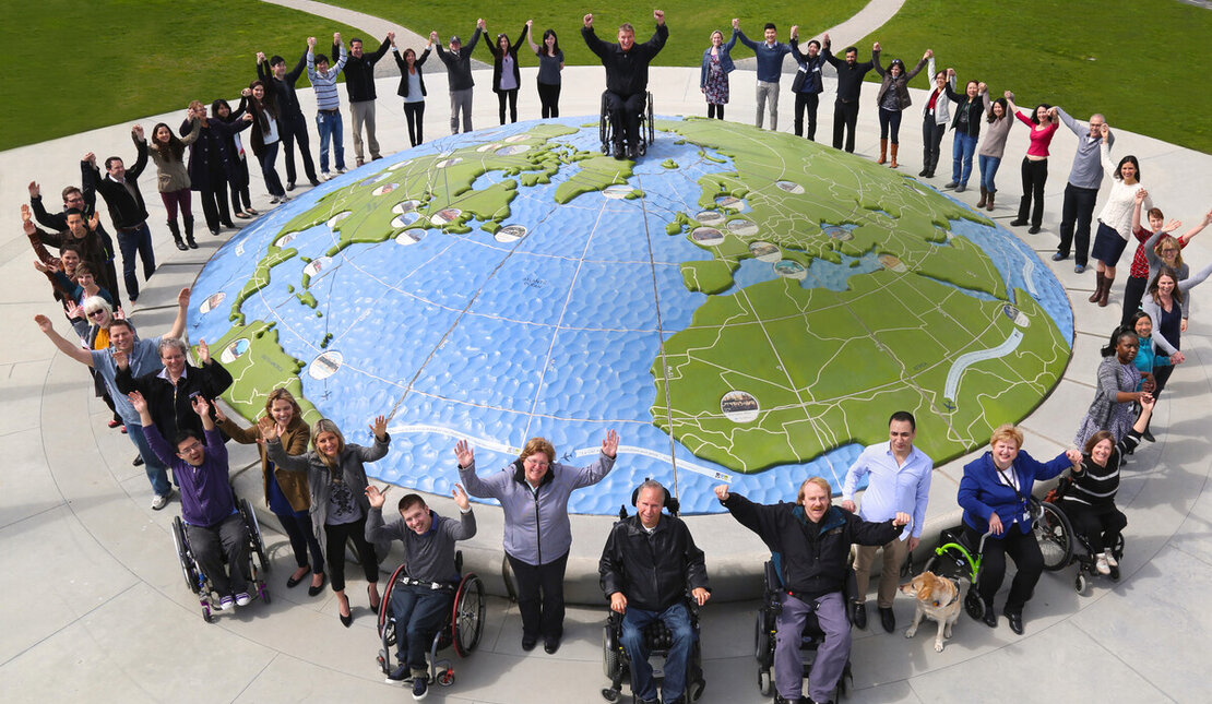 A group of people of different abilities around the perimeter of a large globe. They are celebrating. 