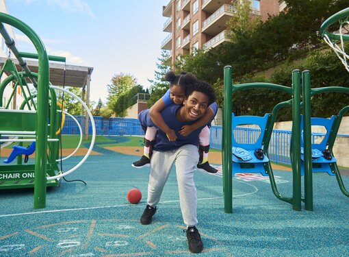 Black man and Black little girl in an accessible playground. The man has the little girl on his back and they are smiling.