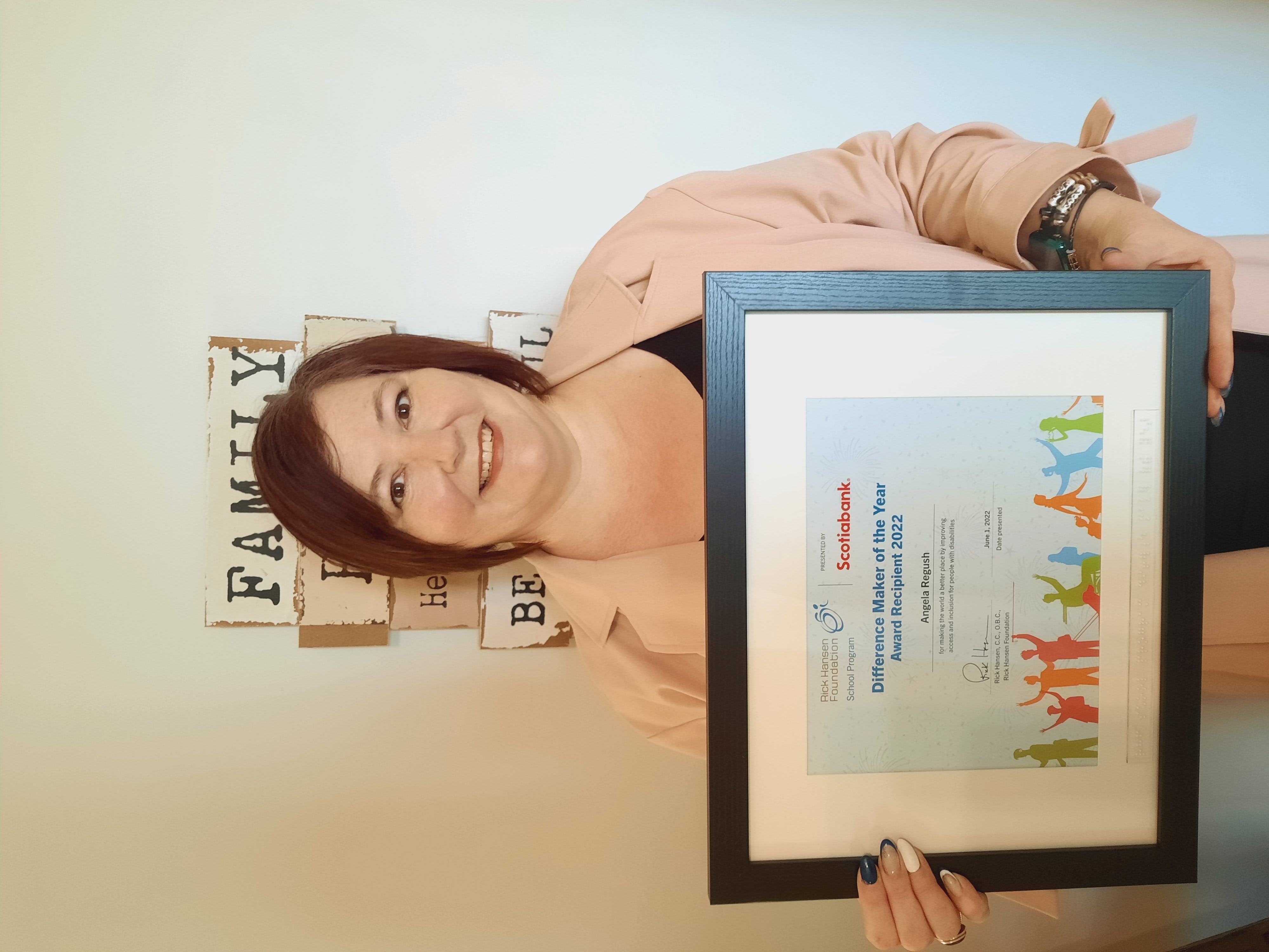 Woman with short brown hair, smiling, holding a certificate proudly in front of wall art that says 'Family'