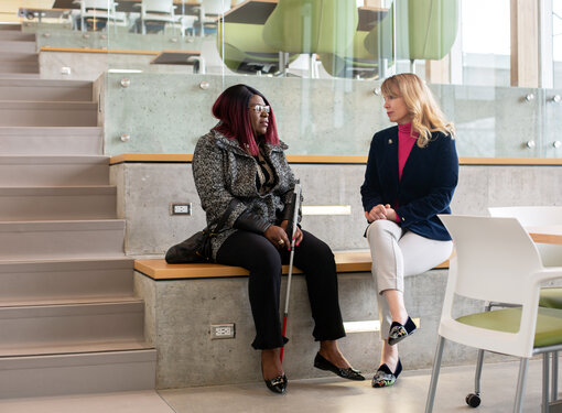 Laetitia Mfamobani, who has long pink and black hair and is wearing a white and black coat sitting beside Rebecca Blissett, who has long blonde hair and is wearing a pink shirt and navy blazer. The two are sitting on stair seats, Laetitia has a white cane.