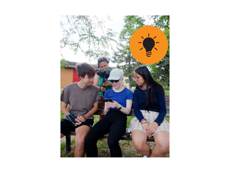 Three teenager siblings sitting on a park bench. On the left is a teenage boy, in the middle is a teenage girl with white hair wearing sunglasses and using a white cane, and on the right is a teenage girl with long black hair.