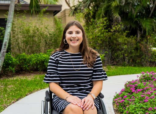 Girl with long brown hair sitting in a wheelchair outdoors, smiling and wearing a blue dress