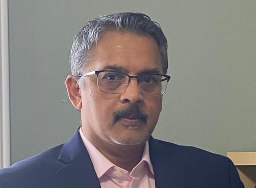 Person with short grey hair and a dark moustache wearing glasses, a pink button up shirt and a dark jacket.