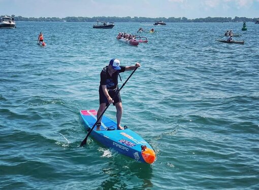 Mike Shoreman paddleboarding. He is wearing a black t-shirt and shorts and a Toronto Blue Jays baseball cap. His paddleboard is blue and orange. There are boats behind him.