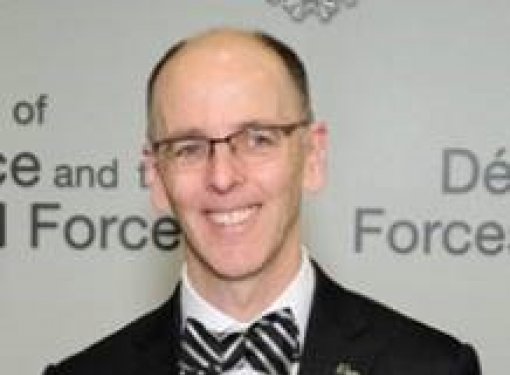 Middle aged man with short hair, glasses, smiling, grey suit with bowtie. 